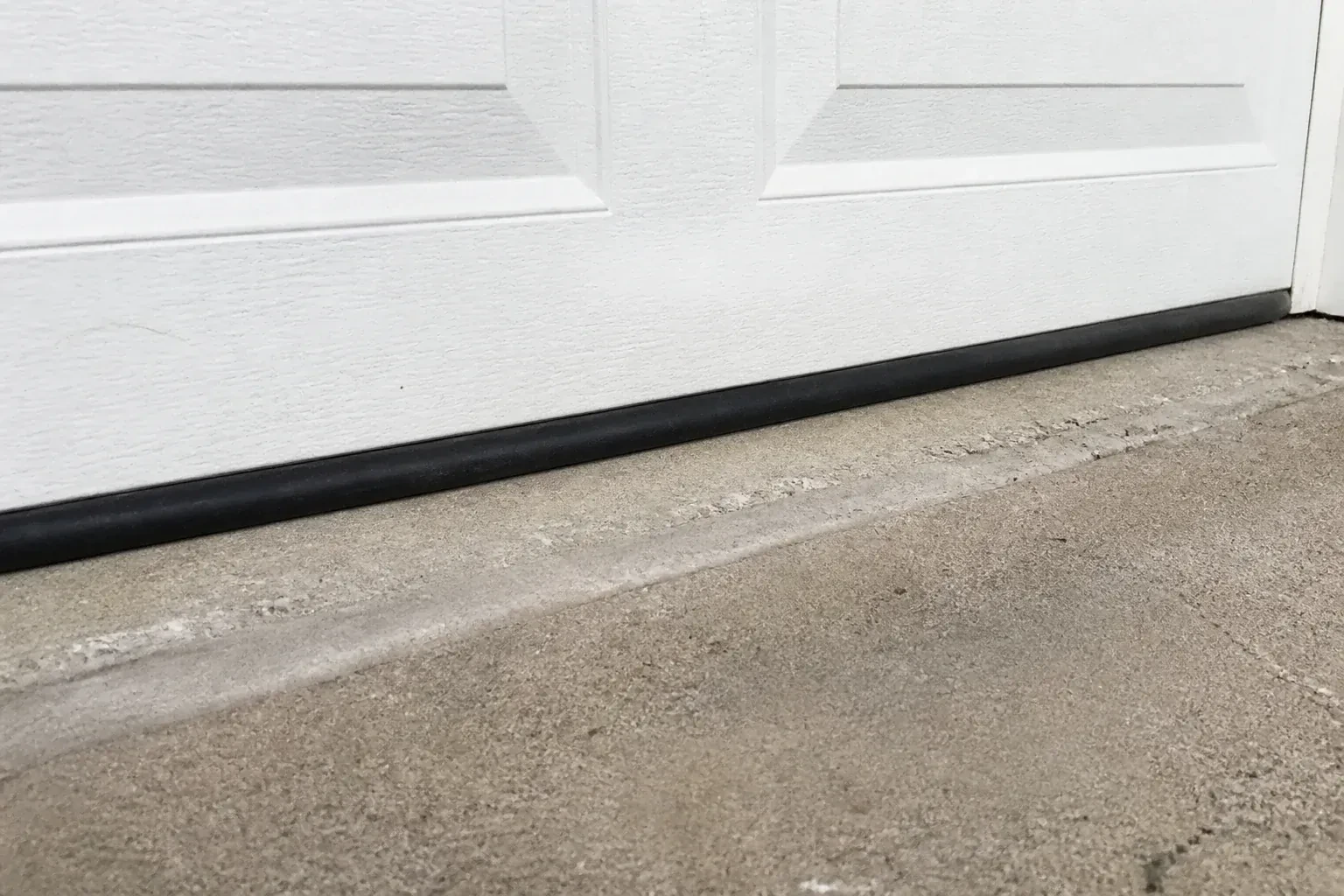 Black bottom seal along a closed white garage door at a concrete threshold. — Garage Door Bottom Seal Replacement, Shelburne